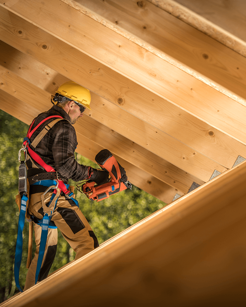 Charpentier travaillant sur une structure en bois avec harnais de sécurité et cloueuse pneumatique.